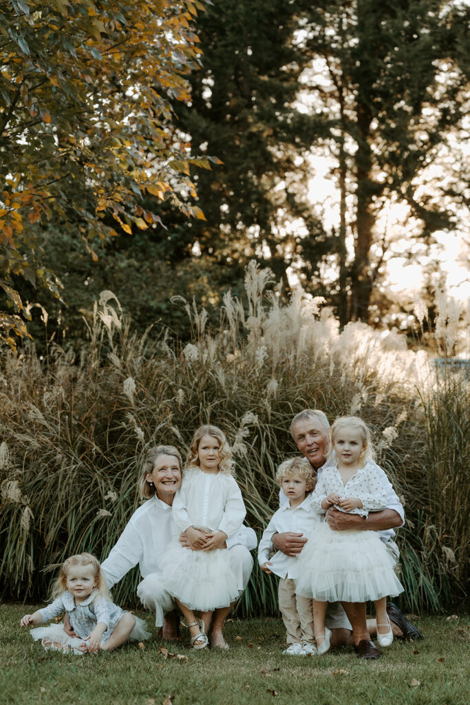 Grandparents kneeling with five grandchildren in a garden, all dressed in white among tall ornamental grasses at sunset.