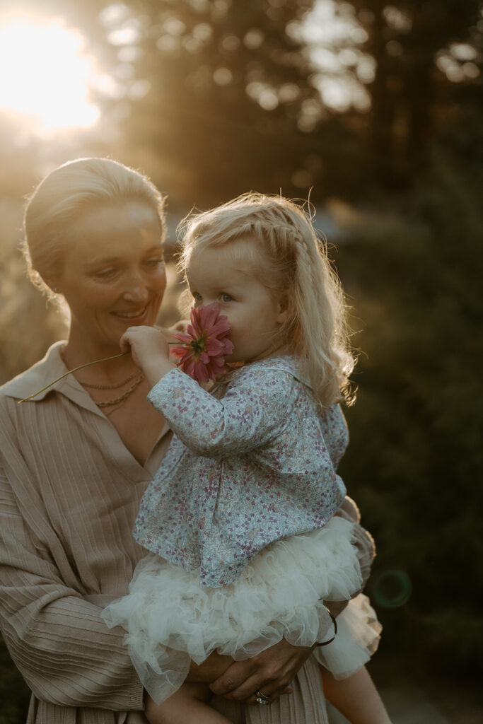 Older woman holding a young girl outdoors at sunset while the girl smells a pink flower.