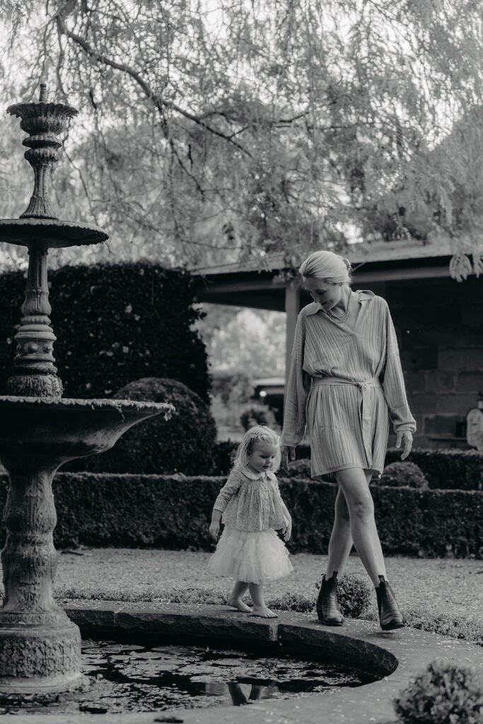 Mother and young daughter walk barefoot along a stone edge by a ornate fountain in a garden; both look down at the water. The scene is in black and white.