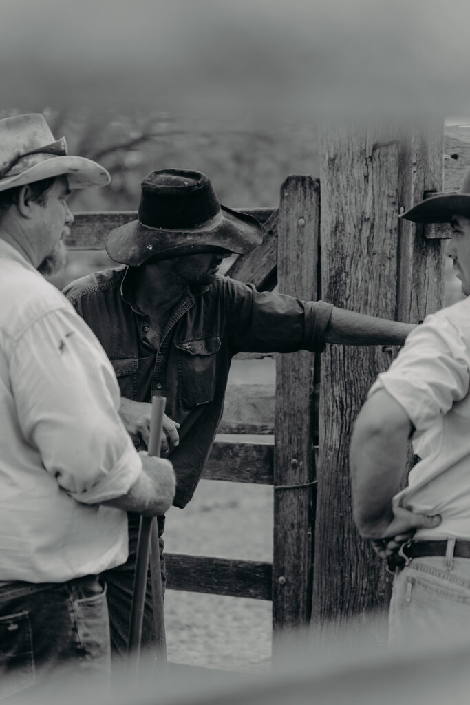 Three men in cowboy hats stand near a weathered wooden fence, one leaning on it and talking with coworkers in a rural setting (black and white).
